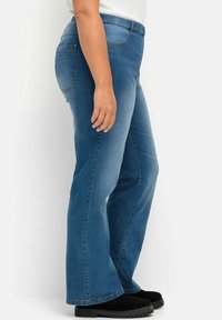 Jeans de mezclilla azul claro con un diseño de pierna acampanada, cinturilla elástica y dos bolsillos traseros. Llevados con botas negras hasta el tobillo.