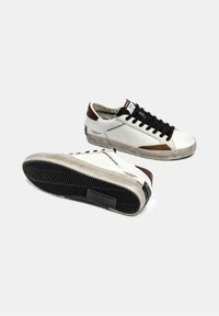 Baskets en cuir blanc avec des lacets noirs, accents de talon marron et une semelle en caoutchouc texturée avec un motif en zigzag. Design décontracté et athlétique.