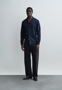 Mann steht vor einer schlichten Wand, trägt ein navyblaues Hemd mit Knöpfen, schwarze weit geschnittene Jeans und schwarze Schuhe, mit neutralem Gesichtsausdruck.