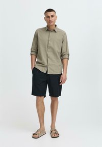 Lys beige lin skjorte med knapfront, rullede ærmer; parret med sorte shorts og brune sandaler, stående på en neutral baggrund.