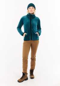 Tamsiai mėlyna šviesi striukė su aukšta apykakle ir juodu užtrauktuku, užsidėta ant rusvų kelnių ir rudos spalvos žygių batų. Modelis stovi su rankomis kišenėse.