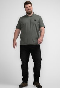 Groene polo met kraag en zwart logo-embleem, gecombineerd met zwarte jeans en bruine schoenen, staande op een neutrale achtergrond.
