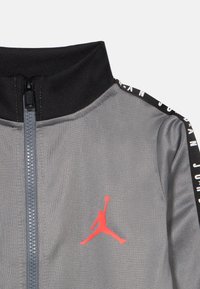Γκρι αθλητικό μπουφάν με μαύρο γιακά, μπροστινό φερμουάρ και κόκκινο λογότυπο Jumpman. Οι πλαϊνές λεπτομέρειες έχουν επαναλαμβανόμενο μαύρο και λευκό κείμενο «JORDAN».