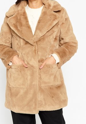 Femme portant un manteau en fausse fourrure beige avec un large col et des poches avant, les mains placées à l'intérieur des poches, par-dessus un pull blanc et un pantalon noir.