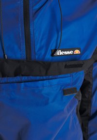 Blue jacket with black accents, featuring a high collar, zippered front, and visible logo. Lightweight, glossy fabric with pockets and Velcro closures.