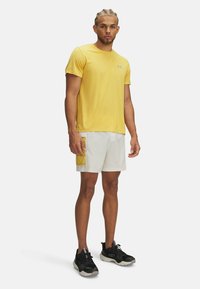 Gelbes, feuchtigkeitsableitendes T-Shirt mit kurzen Ärmeln, kombiniert mit weißen Shorts, die eine gelbe Tasche haben. Schwarze Sportliche Schuhe mit strukturierten Details.