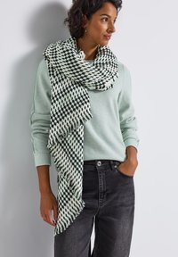 Lysegrøn sweater med lang halvmønstret tørklæde i mørkegrøn og creme. Tørklædet har frynset kanter. Grå jeans.