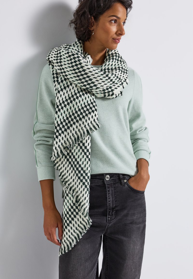 Lysegrøn sweater med lang halvmønstret tørklæde i mørkegrøn og creme. Tørklædet har frynset kanter. Grå jeans.