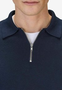 Marinblå zip-up polo med ribbad textur, med krage och silverdragkedjedetalj vid halsringningen över en vit T-shirt.