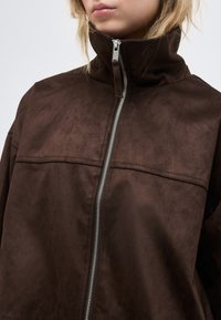 Νεαρό άτομο που φοράει ένα σκούρο καφέ δερμάτινο μπουφάν από σουέντ με ψηλό γιακά και ασημένιο φερμουάρ στο μπροστινό μέρος που είναι μερικώς κλειστό.