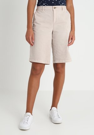 Shorts en coton beige avec une coupe droite, une longueur au genou, des poches avant et une fermeture à bouton, associés à des baskets blanches.