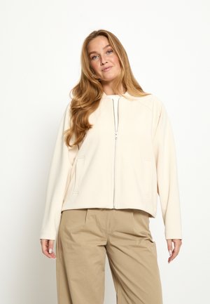 Mladá žena s dlouhými světle hnědými vlasy, oblečená v béžové bundě na zip a hnědých širokých kalhotách, stojící před bílým pozadím.