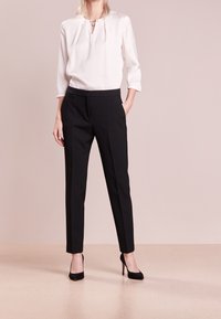 Blouse blanche à manches longues avec un décolleté en V, associée à un pantalon noir ajusté et des escarpins noirs sur un fond clair.