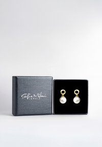 Guldtonade örhängen med runda vita pärlor, presenterade i en mörkgrå texturerad låda med svart inredning och logotyp på toppen.