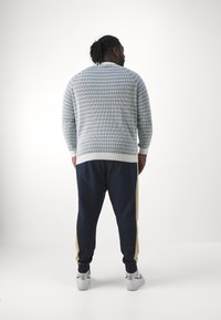 Suéter listrado em azul claro e branco, com punhos canelados, combinado com calças de jogging azuis-marinho com riscas douradas laterais e tênis brancos.