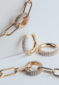 Gylden kædeled med en tekstureret, udsmykket oval, to cirkulære ringe udsmykket med klare ædelsten, glatte og blanke overflader.