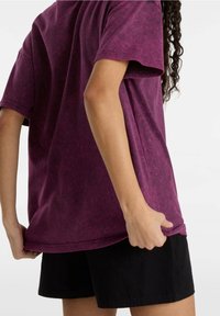 Camiseta de tie-dye burdeos con mangas cortas, que presenta un corte holgado y una textura suave. Combinada con pantalones cortos negros para un look casual.
