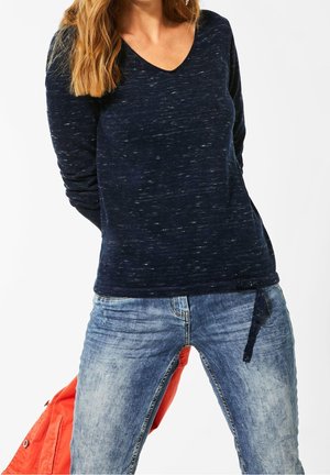 Femme portant un haut à manches longues bleu foncé avec un détail noué sur le côté et un jean bleu clair délavé, tenant une veste orange dans son dos.