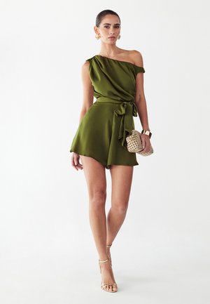 Naine, kes kannab oliivrohelist ühe õlaga kleiti, millel on seotud vöökoht, hoiab kootud kotti ja kannab kuldseid kõrvarõngaid, käevõru ja kontsaga kingi.