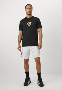 Camiseta negra con un gráfico circular en azul y oro, combinada con pantalones cortos de malla blancos con franjas laterales negras, y zapatillas negras.