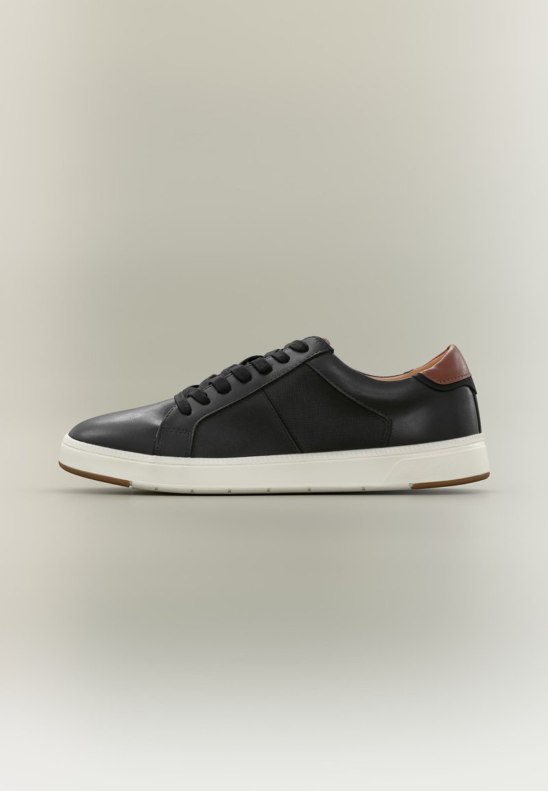 Μαύρο sneaker χαμηλής κορυφής με καφέ ταμπελάκι στη φτέρνα, λευκή σόλα και μαύρα κορδόνια σε ουδέτερο φόντο.