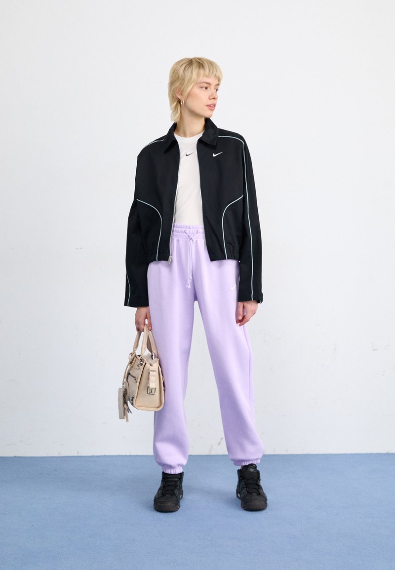 Chaqueta corta negra con acentos blancos, camiseta blanca, pantalones de chándal lavanda y zapatillas negras. Sosteniendo un bolso ligero color beige.