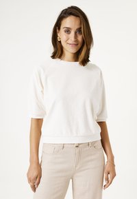 Weißer Baumwollpullover mit rundem Halsausschnitt und kurzen, hochgerollten Ärmeln. Der Schnitt ist locker, und er hat einen gerippten Saum und Bündchen. Getragen mit beigen Hosen.