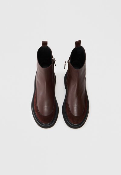 Een paar bruine leren enkelboots met zwarte chunky zolen en zijritsen, afgebeeld vanuit een bovenaanzicht op een witte achtergrond.