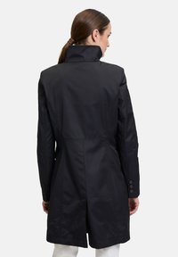 Schwarzer Trenchcoat mit hohem Kragen, taillierter Passform, subtiler Textur und knöpfbaren Ärmeln. Verfügt über einen rückwärtigen Schlitz für mehr Bewegungsfreiheit.