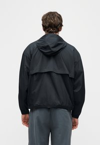 Άνδρας με φοράει μαύρο αδιάβροχο με κουκούλα και σκουρόχρωμο γκρι παντελόνι, στραμμένος προς τα πίσω, με λευκό απλό φόντο.