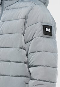 Grå pufferjakke med et quiltet design, der har vandrette sømme, en glat tekstur og et genkendeligt sort mærke på venstre ærme.