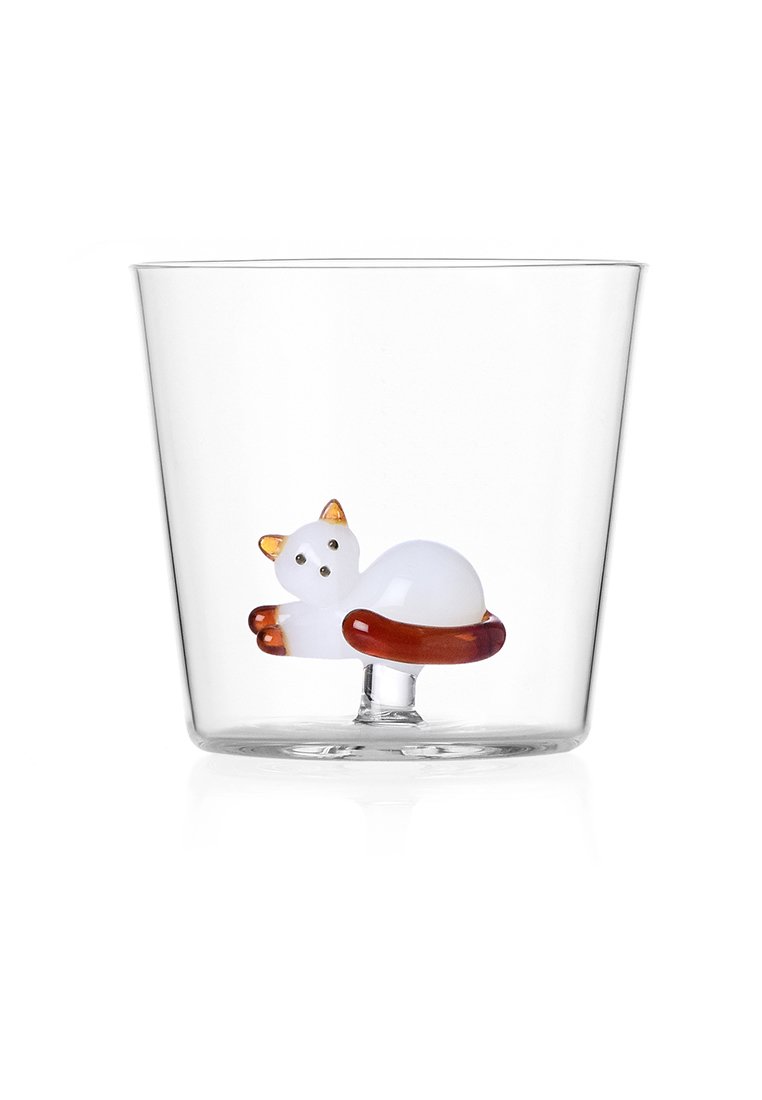 Przezroczysta szklanka z białą figurką kota z pomarańczowymi akcentami, spoczywająca na brązowej okrągłej podstawce wewnątrz kubka.