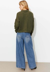 Pull en maille épaisse vert olive avec ourlet côtelé, associé à un jean bleu clair à jambes larges. Des chaussures noires à talons hauts complètent la tenue.