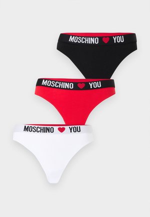 Noor naine seisab paljajalu mustas Moschino bralette'is ja sobivate pükstega, mille vöökohal on südame detail, ühevärvilise valge taustaga.