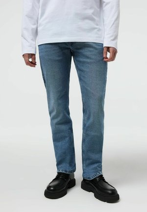 Mężczyzna noszący jasnoniebieskie obcisłe dżinsy, czarne masywne sznurowane buty oraz białą koszulę z długim rękawem, stojący na białym tle.