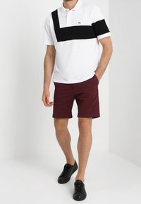 Bílá polokošile s černým diagonálním pruhem, doplněná o bordó kraťasy. Černé tenisky dotvářejí outfit a zdůrazňují ležérní styl.