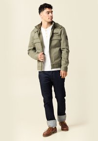 Man in een olijfgroen jack, witte henley shirt, donkere opgerolde jeans en bruine leren laarzen die tegen een neutrale achtergrond staat.