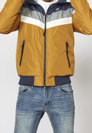 Person trägt eine senfgelbe, graue und marineblaue Jacke mit Reißverschluss und blaue verwaschene Jeans, Arme über dem Kopf außerhalb des Bildausschnitts erhoben.