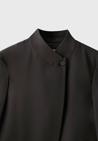 Veste noire avec un col mandarin haut et un seul bouton visible près de l'encolure sur un fond uni.