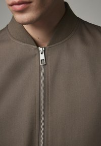Braune Bomberjacke aus strukturiertem Stoff, mit rippendem Kragen und silbernem Reißverschluss. Das Design zeigt eine Nahaufnahme des Ausschnitts und des Reißverschlusses.