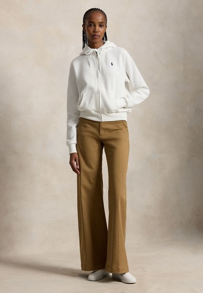 Woman with braided hair wearing a white zip-up hoodie, tan wide-leg pants, and white shoes standing against a neutral background.