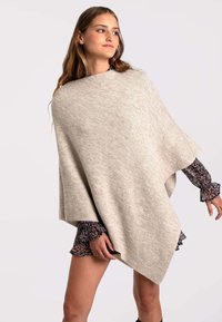 Poncho de punto beige con un diseño drapeado y una textura suave, llevado sobre un vestido estampado con mangas con volantes. Los bordes son limpios y suaves.