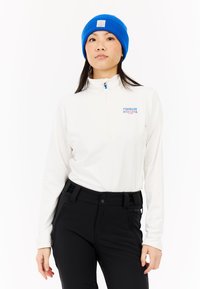 Hvid lynlåset langærmet top med blå lynlås og logo, parret med sorte bukser. Modellen bærer en blå strikkebeanied. Enkel, funktionel design.