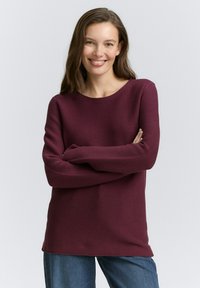 Burgunderfarbener Rippstrickpullover mit rundem Halsausschnitt und langen Ärmeln, kombiniert mit blauen Jeans. Glatte Textur, lockere Passform.