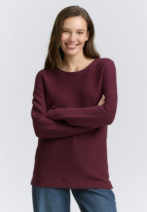Pull-over côtelé bordeaux avec un col rond et des manches longues, associé à un jean bleu. Texture douce, coupe décontractée.
