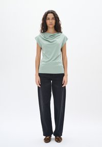 Top drappeggiato verde menta con maniche a cappuccio, abbinato a jeans neri larghi e scarpe leopardate, su uno sfondo bianco semplice.