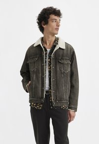 Denim jacket with a beige fur collar, featuring two chest pockets, dark wash, and button front. Worn over a patterned black shirt.