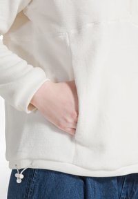 Sudadera de forro polar blanca con una textura suave, cordón ajustable en el dobladillo y un bolsillo frontal. Los puños son acanalados y ajustados.