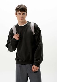 Svart oversized sweatshirt med ribbad krage och muddar, tillsammans med grå byxor. Modellen bär en ljusgrå ryggsäck.