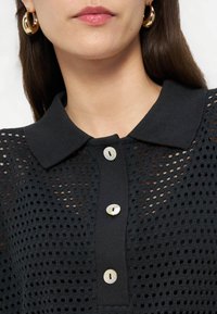 Γυναίκα που φοράει μαύρο πλεκτό τοπ με γιακά και κουμπιά από φίλντισι καθώς και χρυσούς κρίκους στα σκουλαρίκια, με τα χείλη της να έχουν διακριτικό ροζ γυαλιστερό κραγιόν.